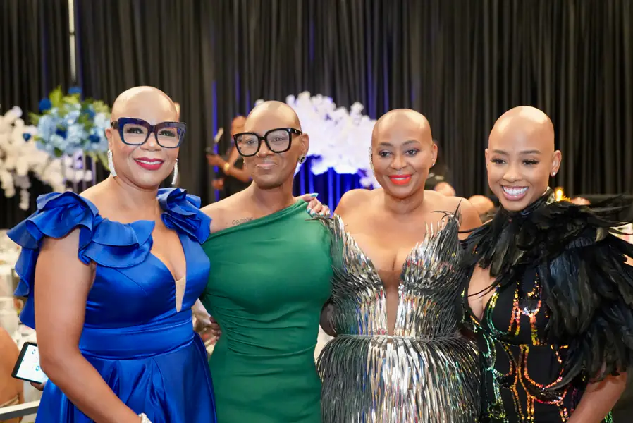 Four bald people pose for a photo in their elegant formal dresses colored blue, green, silver, and black.