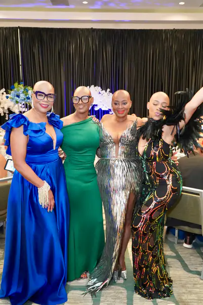 Four bald people pose together at a conference while wearing formal dresses in blue, green, silver fringe, and a black sequined style with feathers.