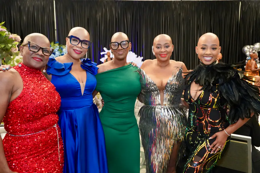 Five bald people stand side by side posing for a photograph while wearing formal evening gowns.