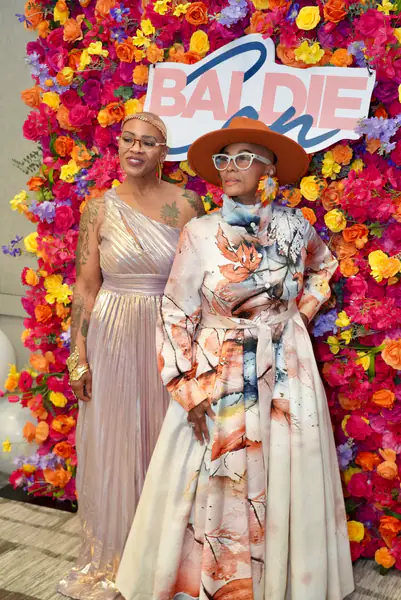 Two bald people stand in front of a floral backdrop as one person wears a metallic gold one-shoulder dress and the other person wears a long leaf-print dress with a wide-brimmed hat.