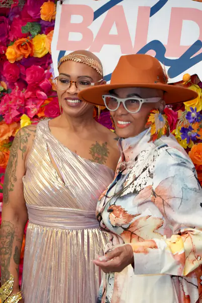Two bald people stand together in front of a floral backdrop as one person wears a gold headband and a metallic dress while the other person wears a rust-colored hat and a patterned shirt.