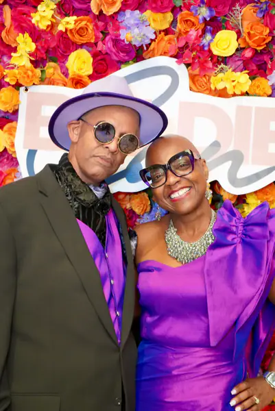 Two bald people pose together in front of a flower wall while one person wears a purple hat, sunglasses, and a suit with a purple vest, and the other person wears dark glasses and a purple one-shoulder dress.