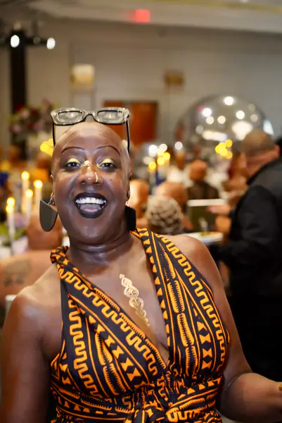 A bald person smiles at the camera while wearing black-rimmed glasses atop their head and a black and orange patterned dress.