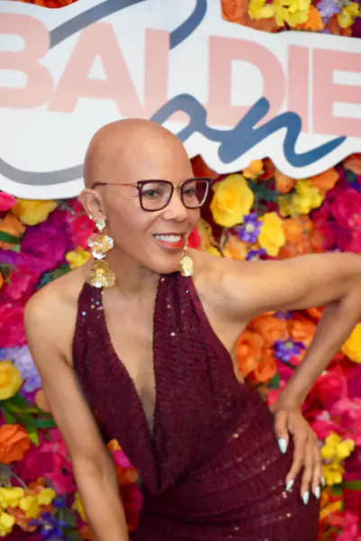 A bald person poses in a maroon halter dress and large gold flower earrings in front of a floral backdrop.