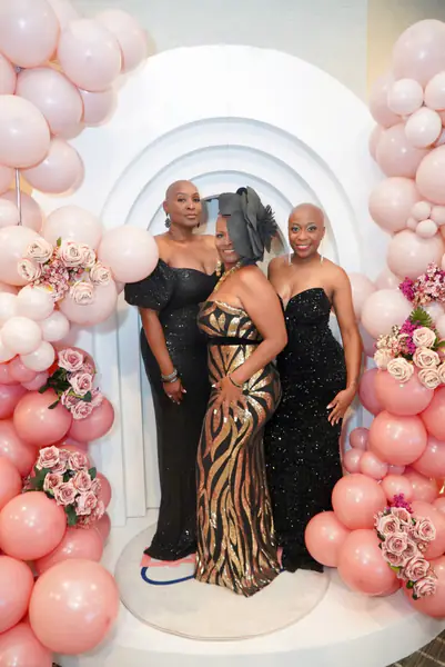 Three bald people pose in elegant floor-length evening gowns in front of an archway decorated with pink balloons and roses.