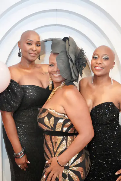 Three bald people pose together while wearing formal black and gold dresses and statement jewelry.