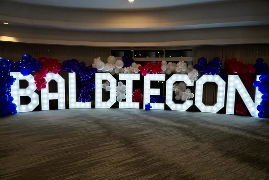 Large light-up marquee letters spelling out BALDIECON stand before a backdrop of red, white, and blue balloons to celebrate a gathering of bald people.