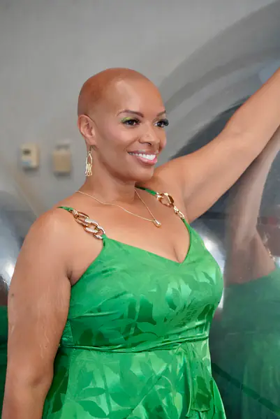 A bald person smiles while leaning against a large reflective surface, wearing a green dress with gold chain straps and gold dangling earrings.