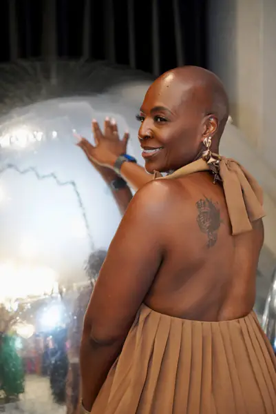 A bald person in a tan halter dress smiles while looking back at a large metallic sphere.