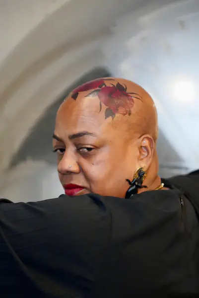 A bald person wearing red lipstick and large figural earrings looks over their shoulder to reveal a colorful rose tattoo on their scalp.