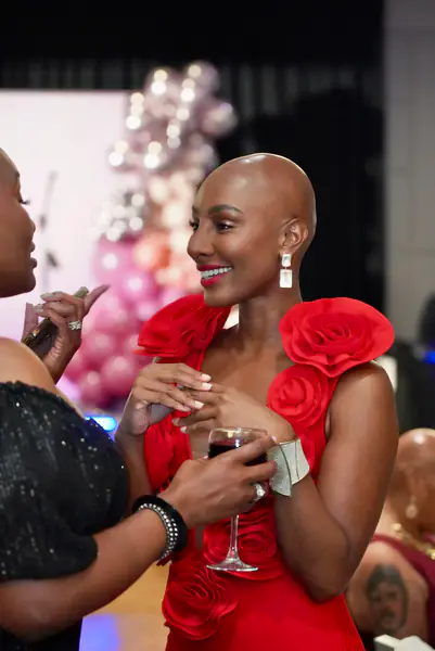 Two bald people talk while holding glasses at a gathering, with one person wearing a bright red dress with large fabric rosettes and the other person wearing a black off-the-shoulder top.