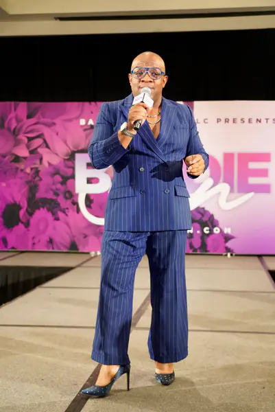 Derek J speaks into a microphone on stage in a blue pinstripe suit and blue-rimmed glasses, in front of a pink backdrop.