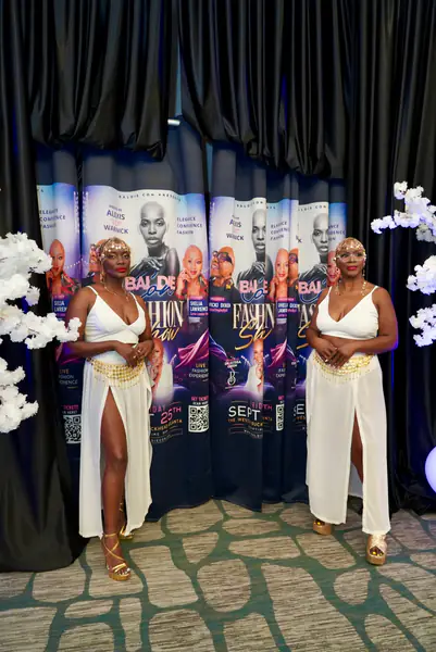 Two models in matching white two-piece outfits with gold waist chains and headpieces pose on stage.