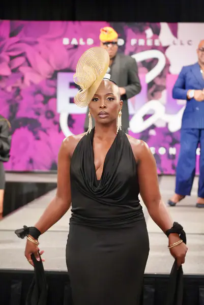 A model walks the runway in a black halter dress, a large gold disc fascinator and pearl drop earrings.