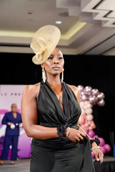A bald model wears a black halter dress, long dangling earrings and a wide-brimmed straw fascinator.