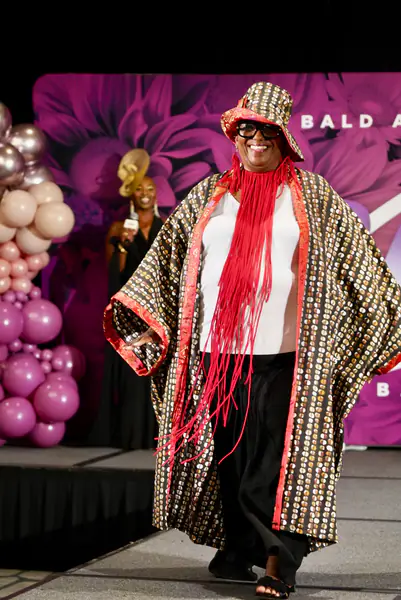 A smiling model walks the runway in a bucket hat, a long red fringe necklace and a patterned open robe.