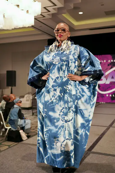 A bald model in oversized glasses walks the runway in a long blue and white tie-dye kaftan with large fringe earrings.