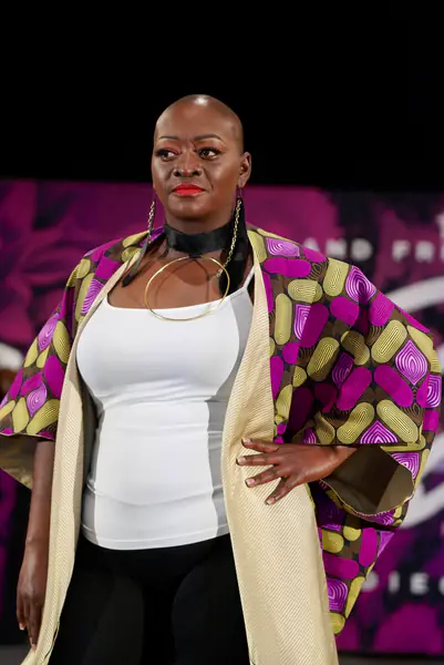 A bald model wears a white tank top, black pants and a wide-sleeved kimono-style jacket with a purple, yellow and black geometric print.