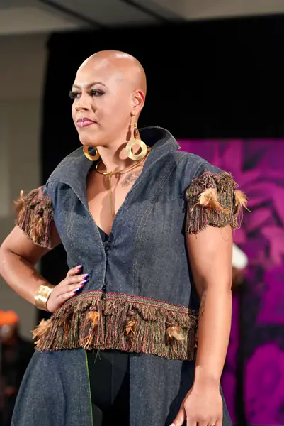 A model wears a denim vest with fringed trim and feather accents, large gold hoop earrings and a gold cuff.