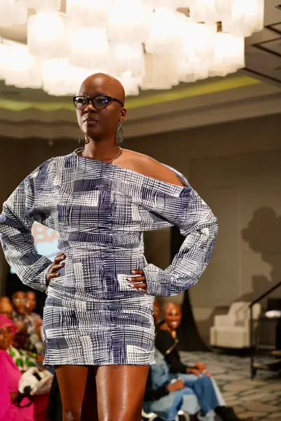 A bald model in glasses walks the runway with hands on hips in a blue and white patterned off-the-shoulder mini dress.