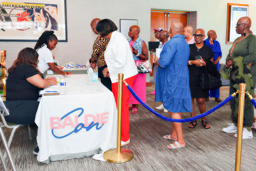 People wait in a line at a registration table while staff help them check in, all standing before a Baldie Con banner.