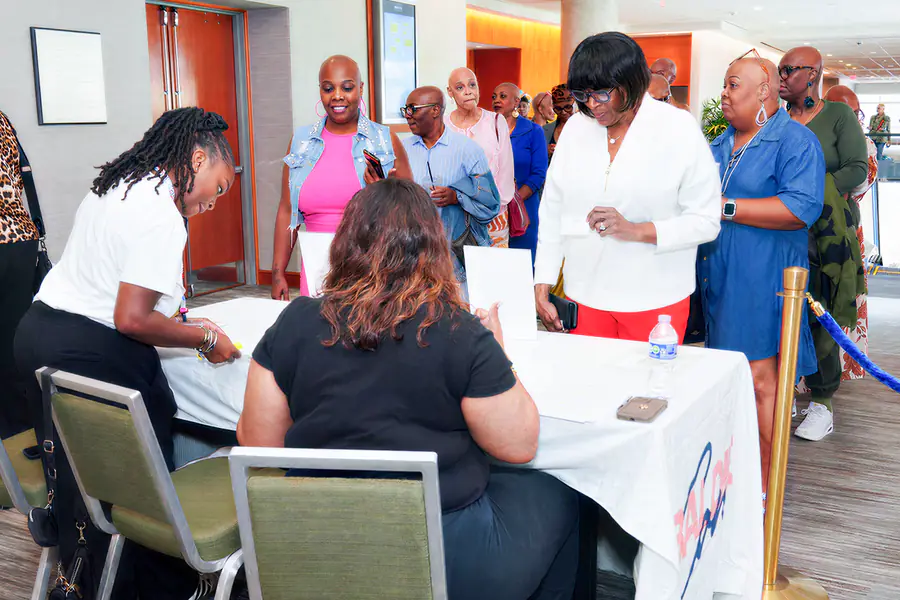 Several bald people wait in a queue at a registration table while two people assist them, and one person wears a white shirt with the word Baldie visible on the table cover.