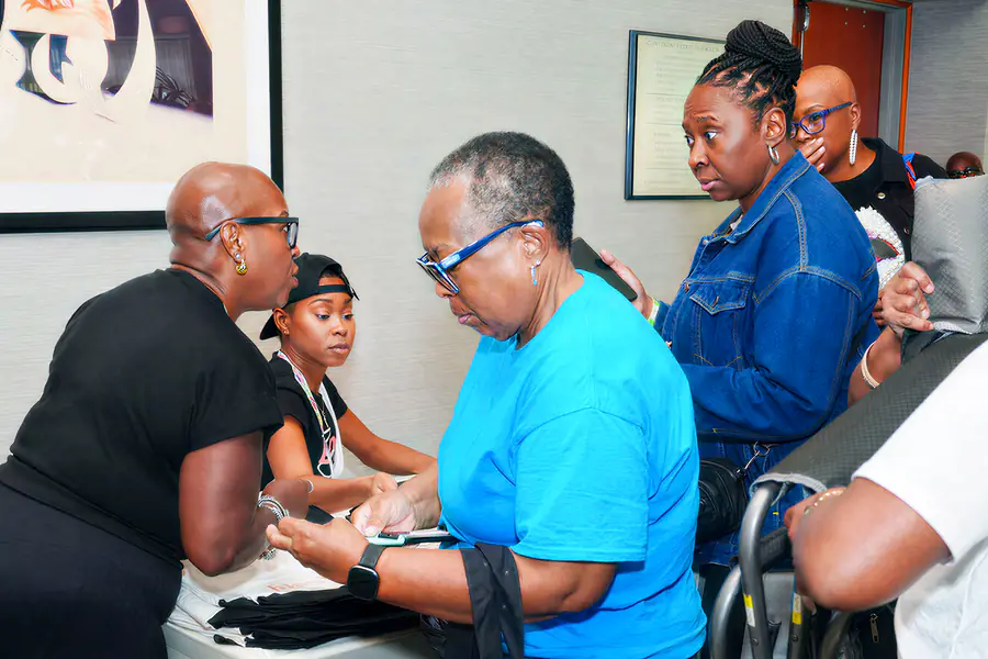 A bald person in a black shirt leans over a table to speak with a person in a blue shirt who is looking at their phone while another person in a denim jacket waits in line behind them.
