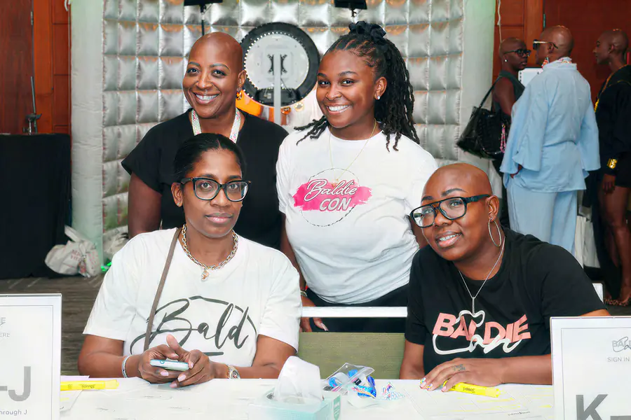 Four people pose together at a registration table, and three of the bald people wear shirts that feature the text Baldie Con.