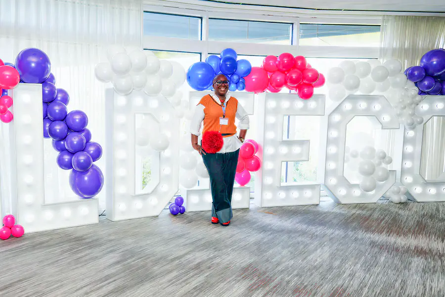A bald person stands with their hands on their hips in front of large light-up letters decorated with purple, pink, and white balloons while wearing an orange sweater vest over a white shirt, dark trousers, and glasses.