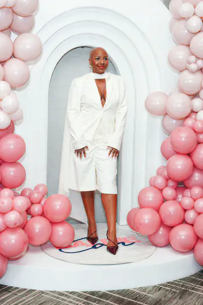 A bald person stands in a white blazer and matching shorts in front of an arched backdrop framed by pink balloons.