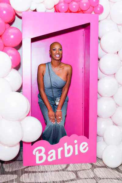 A bald person poses inside a pink Barbie display box while surrounded by pink and white balloons.