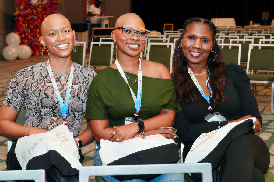 Two bald people and one person with long hair sit together at a conference wearing branded lanyards and holding white tote bags with printed text.