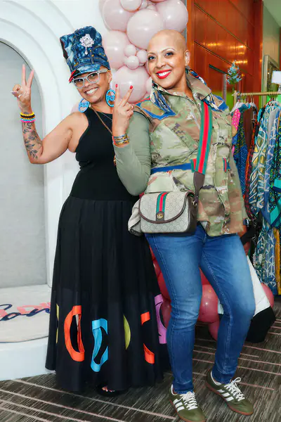 Two bald people pose for a photo with peace signs, one wearing a black maxi skirt with colorful lettering and the other wearing a camouflage jacket and jeans.