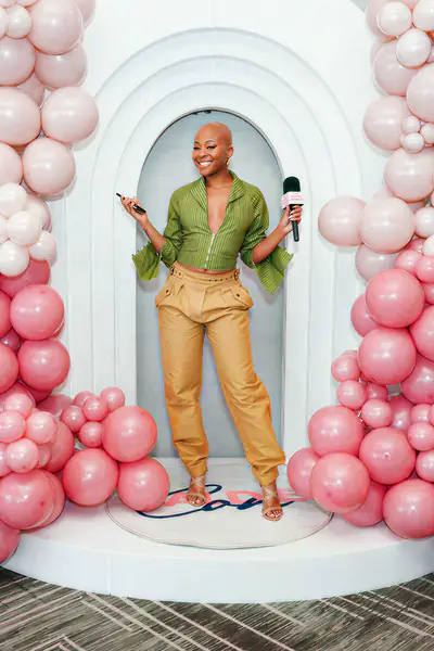 A bald person smiles while holding a microphone and a phone in front of a white arched backdrop decorated with pink balloons while wearing a green zip-up top and tan pants.