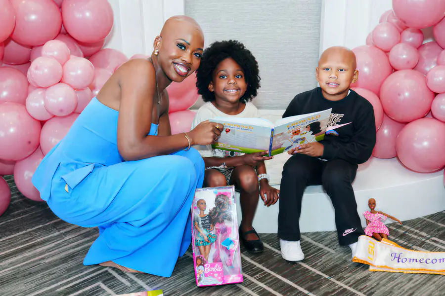 A bald person in a blue outfit holds a book for two children to read while a bald child sits next to them in front of pink balloons.