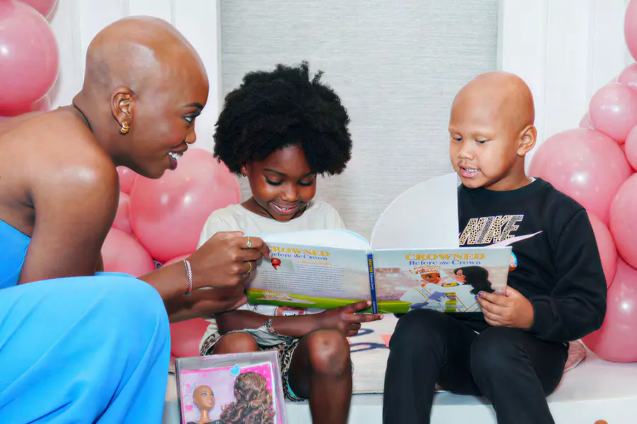 A bald person reads a book titled CROWNED Before the Crown to two children, one of whom is bald and wearing a black Nike sweatshirt.