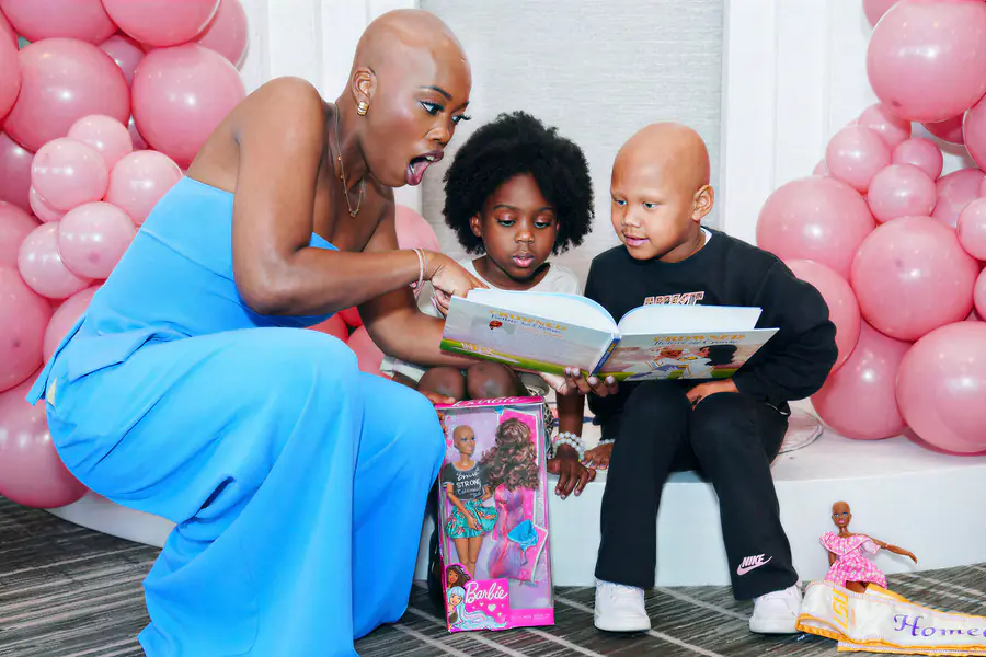 A bald person in a blue jumpsuit reads a book to two children, one of whom is bald, while a bald Barbie doll in a box sits in the foreground.