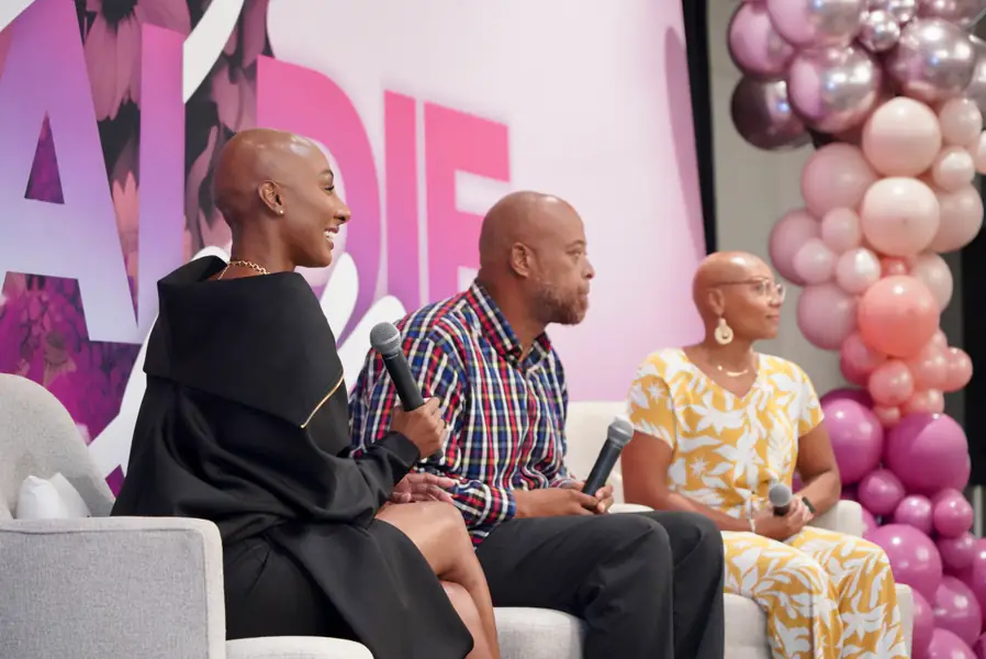 Three bald people sit on a stage in chairs holding microphones while participating in a panel discussion.