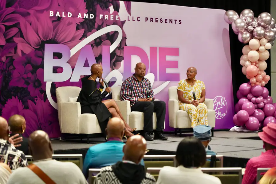 Three bald people sit on chairs during a panel discussion in front of a banner that reads Baldie Con.