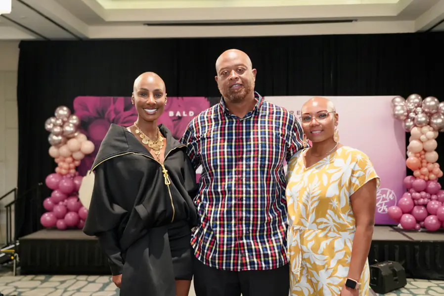 Three bald people stand together smiling at a conference while positioned in front of pink and gold balloon arches.