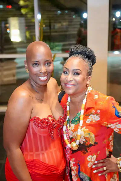 A bald person in a red strapless dress stands with their arm around a person in a bright orange patterned shirt who has their hair in an updo.
