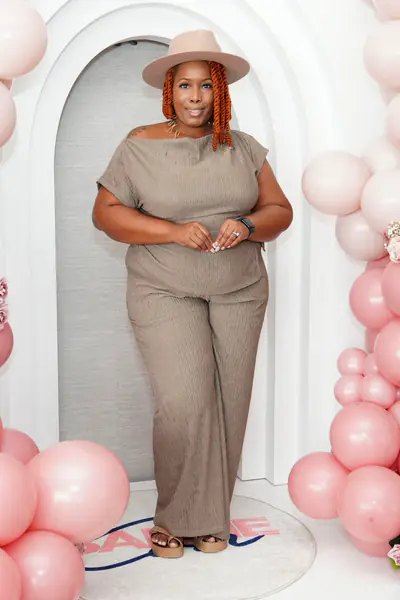 A bald person stands between columns of pink balloons while wearing a tan hat and a textured beige two-piece outfit.