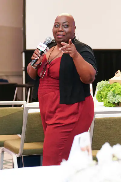 A bald person gestures toward the audience while speaking into a microphone, wearing a red jumpsuit and a black cardigan.