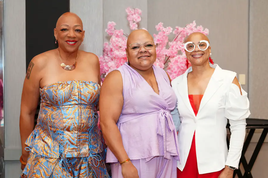 Three bald people stand together smiling, wearing a blue patterned strapless top, a light purple sleeveless top, and a white blazer over a red outfit.