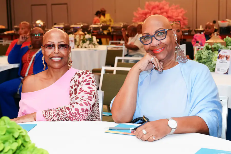 Two bald people sit at a table smiling at the camera as one person wears a pink animal print top and the other person wears a light blue top with large triangular earrings.