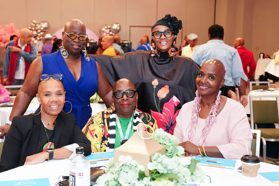 Four bald people pose for a photo at a table while wearing a blue jumpsuit, a floral-patterned black tunic, a black blazer, and a light pink top.