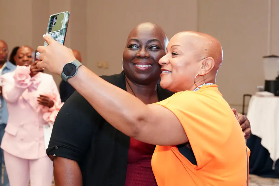 Two bald people smile while taking a selfie together as one person wears a black top and the other person wears an orange shirt.