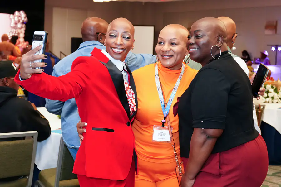 Three bald people pose for a selfie while one person in a red suit holds a phone, another person in an orange top wears a name tag, and a third person wears a black top and maroon pants.