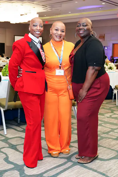 Three bald people pose together while wearing a red suit, an orange jumpsuit, and a black cardigan over a dark red outfit.
