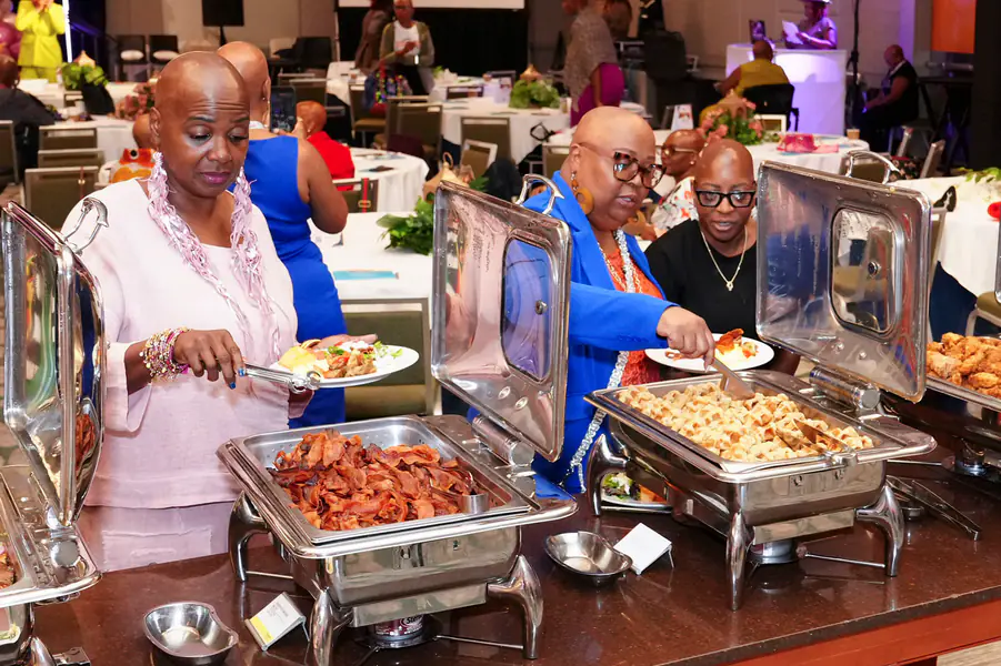 Two bald people serve themselves food from a buffet line while wearing clothing that features text.